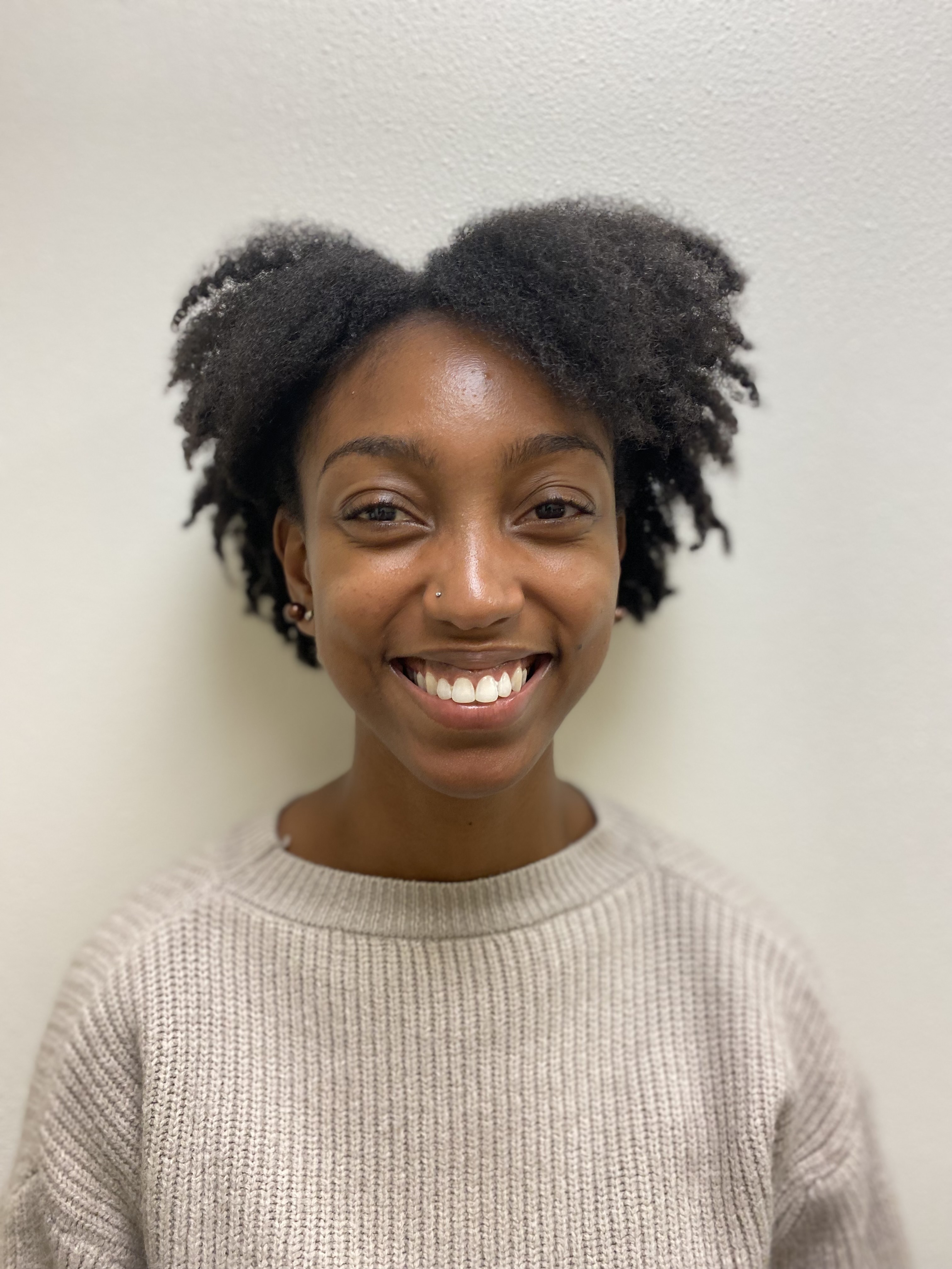 A person (Imani) is in the center of the frame smiling at the camera. She is wearing a knitted, white sweater. She is standing against a blank background.