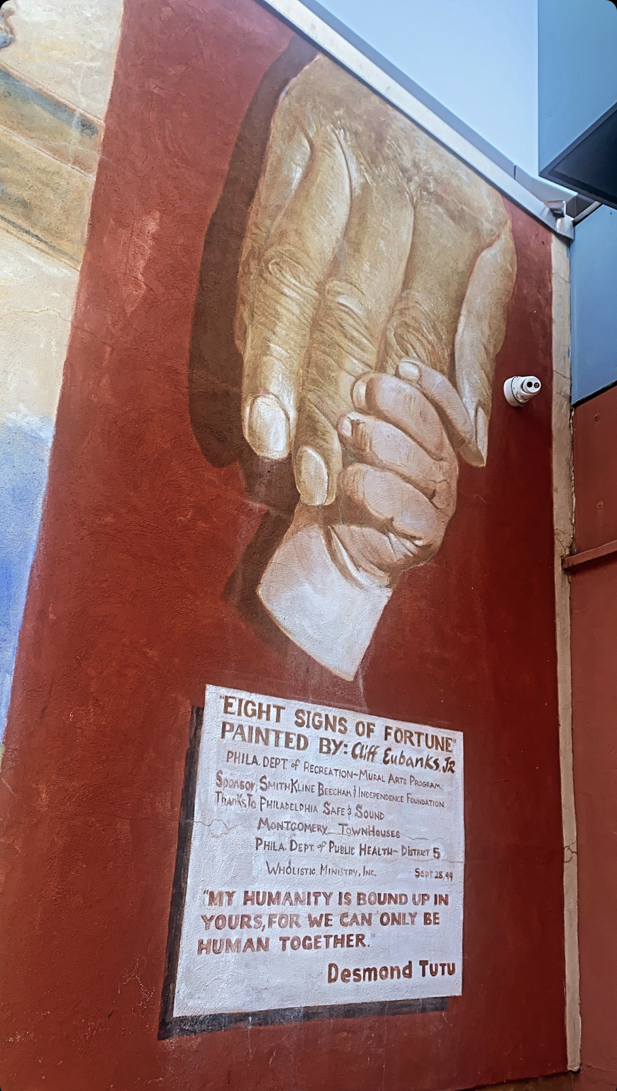 Mural of an adult hand being grasped by a childs hand on a red wall. Under the image is text, some illegible, but the last line is a quote that reads "my humanity is bound up in yours, for we can only be human together" - Desmond Tutu