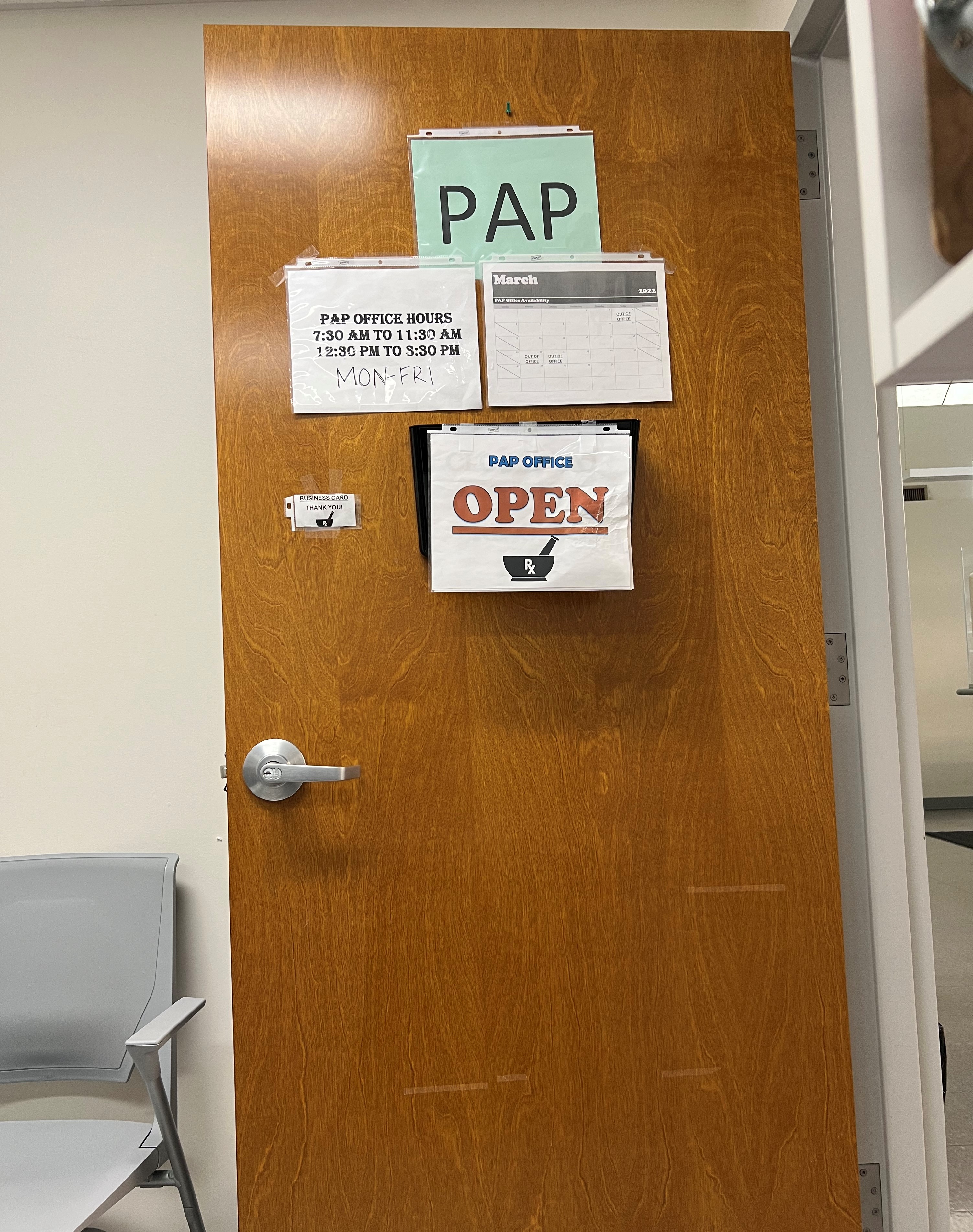 An open door with four signs hanging. At the top a sign reads "PAP" under that are two, on the left lists the office hours and on the right is a calendar showing March. The last sign is below all of them and it reads "PAP Office Open" 