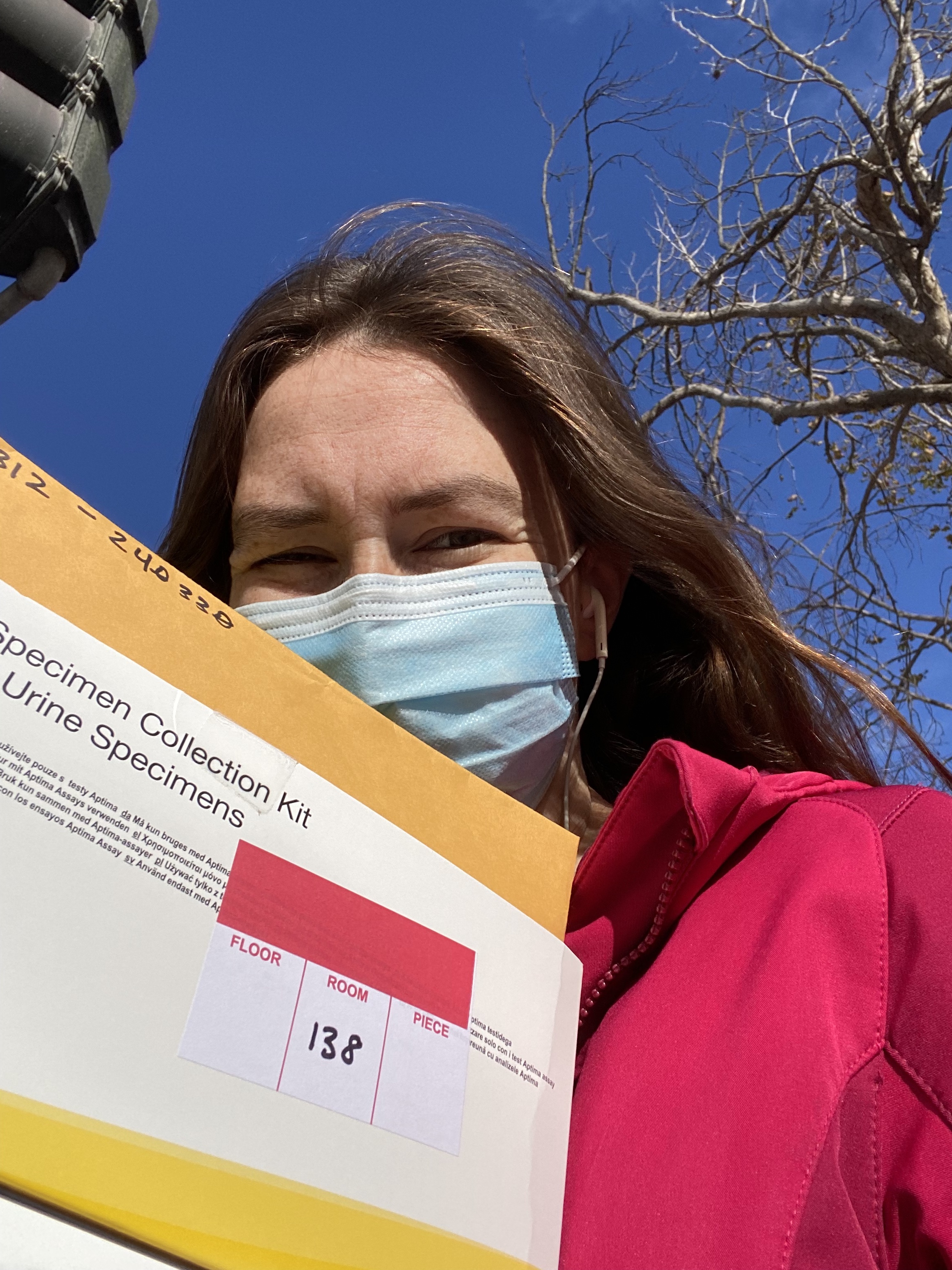 Catherine is seen taking a selfie during service. She is wearing a pink hoodie and a blue medical facemask. She is holding a box labeled "Specimen Collection Kit - Urine Specimens."
