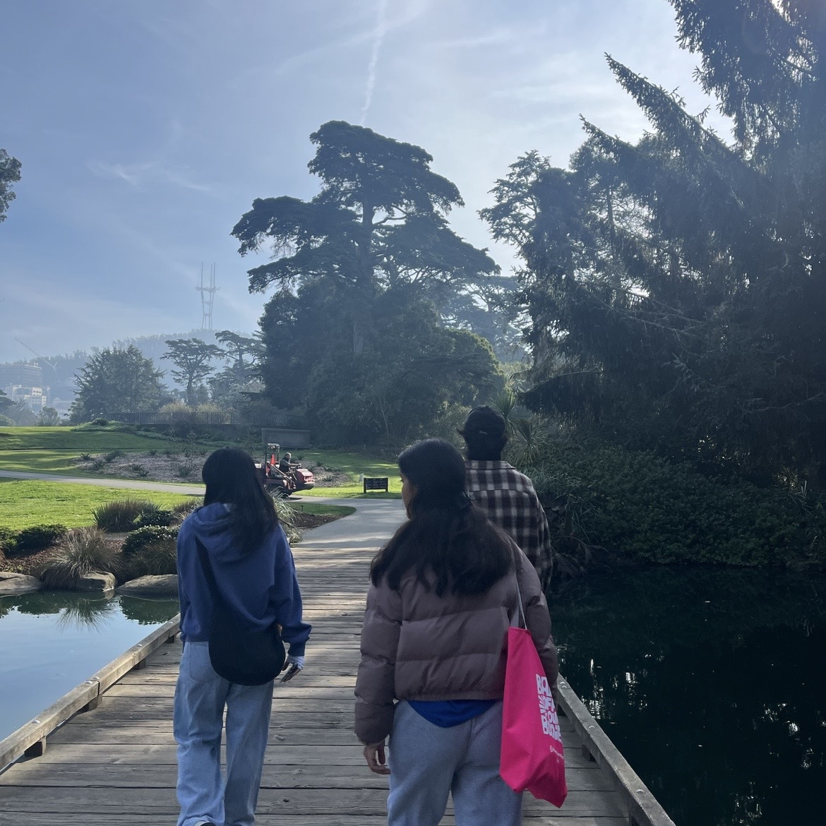 The photo is taken behind a group of 3 NHC members that are walking along a park trail. To the right, there are many trees and bushes, to the left, there is a grassy field. In the distance, a hill is seen behind the light morning fog, with Sutro Tower jutting into the sky.