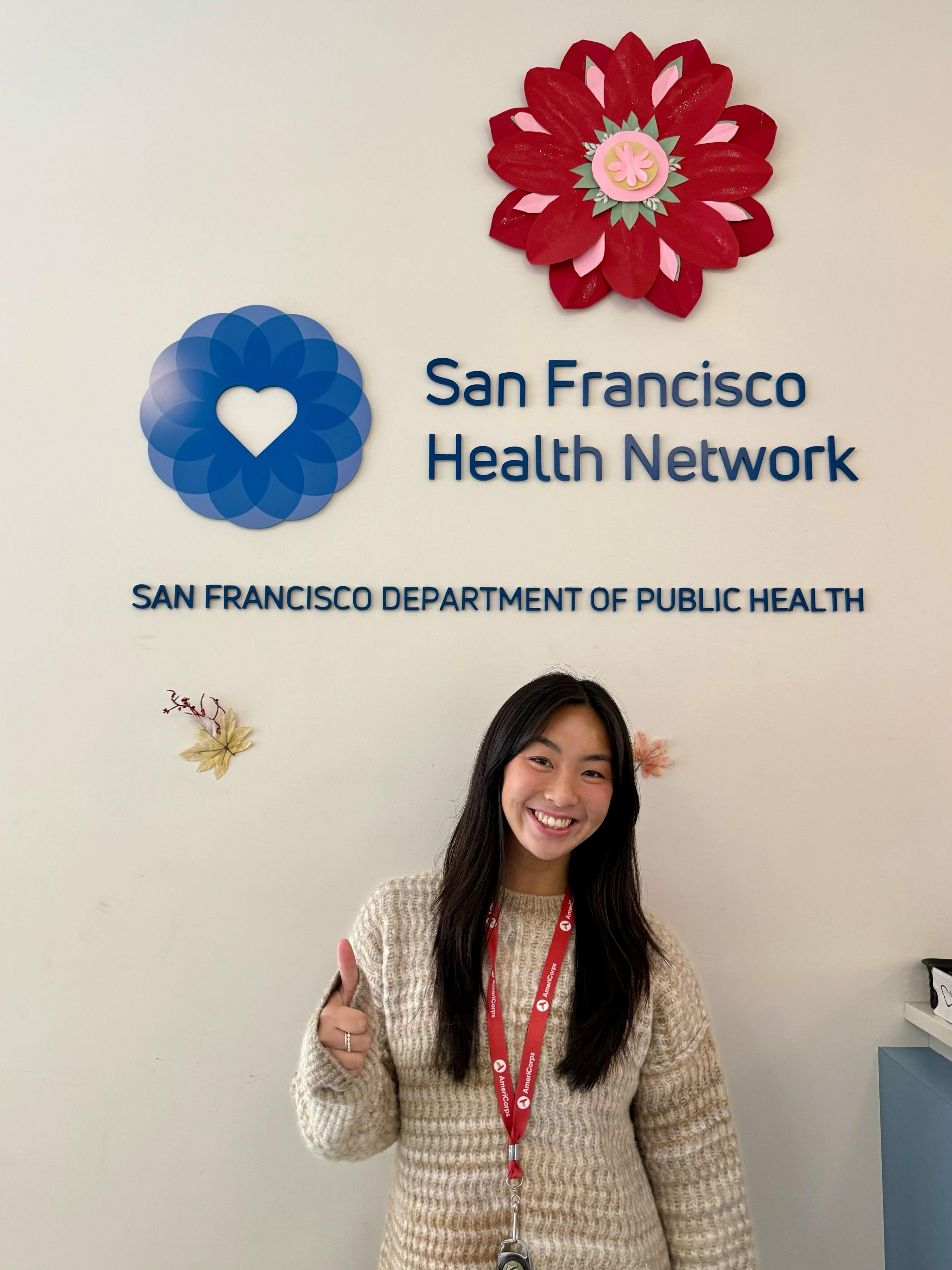 Sam stands in the center of the frame, facing the camera and smiling. Behind her is the logo for the San Francisco Health Network.