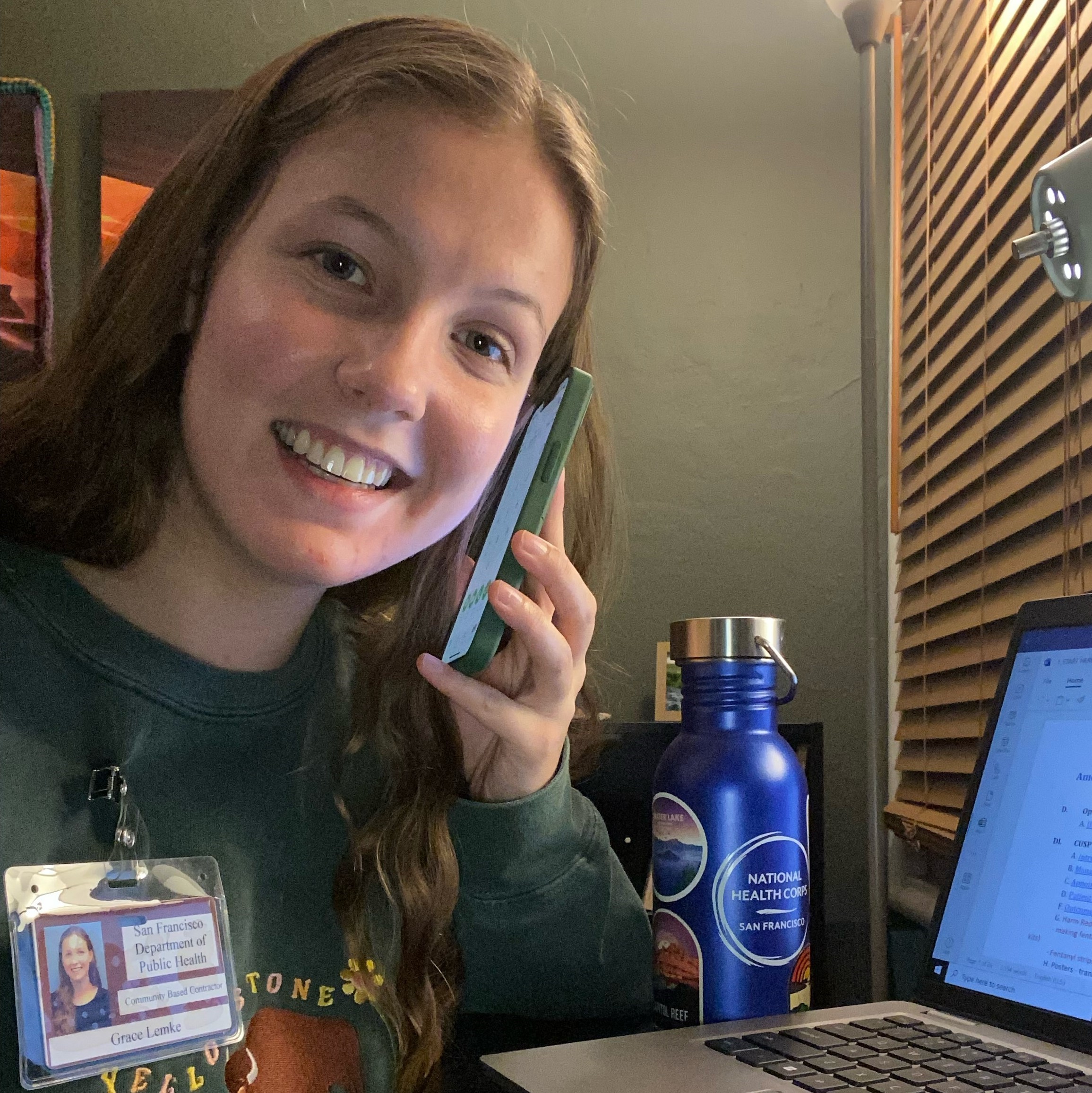 Grace is seen on the phone, while on her computer. In the background is a National Health Corps San Francisco water bottle. 