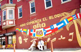 two people sitting in front of a mural on a red brick wall. the mural says "aqui comienza el bloque de oro" which translates to "here begins the block of gold" under the words are flags of many Latin American countries