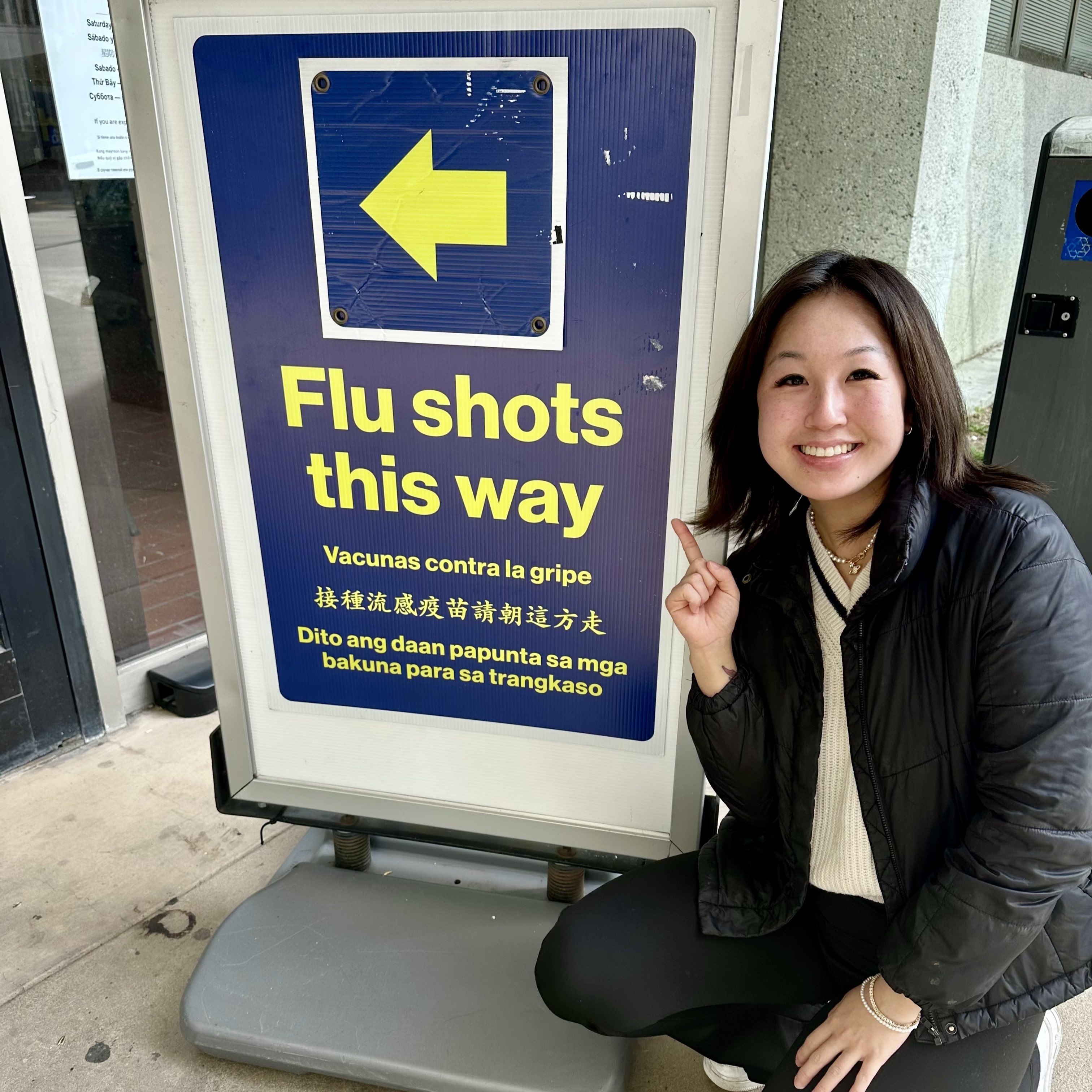 Lilian kneels next to and points towards a sign with an arrow pointing towards the left. On it is written "Flu shots this way" in English, Spanish, Chinese, and Tagalog.