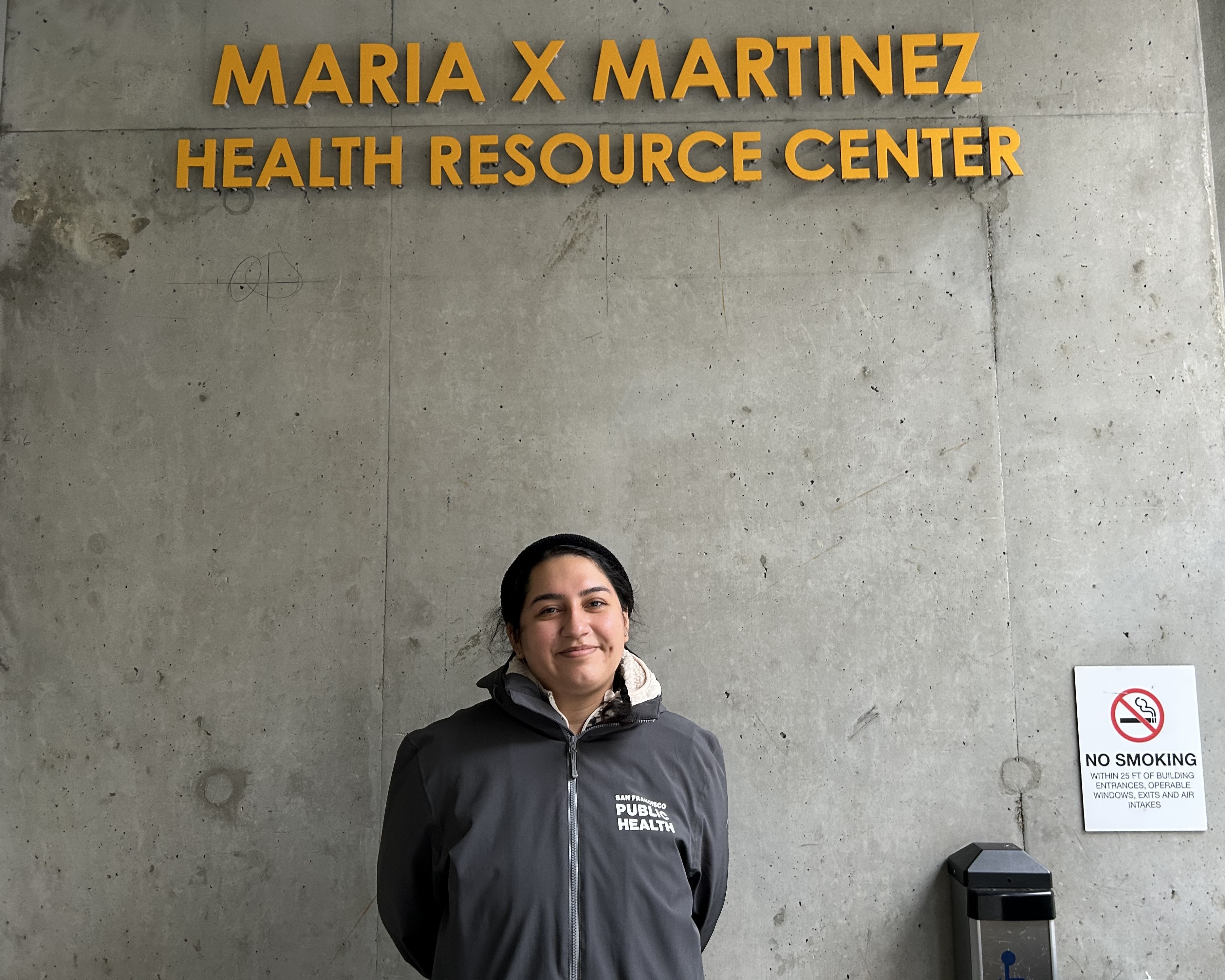 Melanie stands in the middle of the frame, facing the camera and smiling, Above her is the sign that reads Maria X Martinez Health Resource Center. 