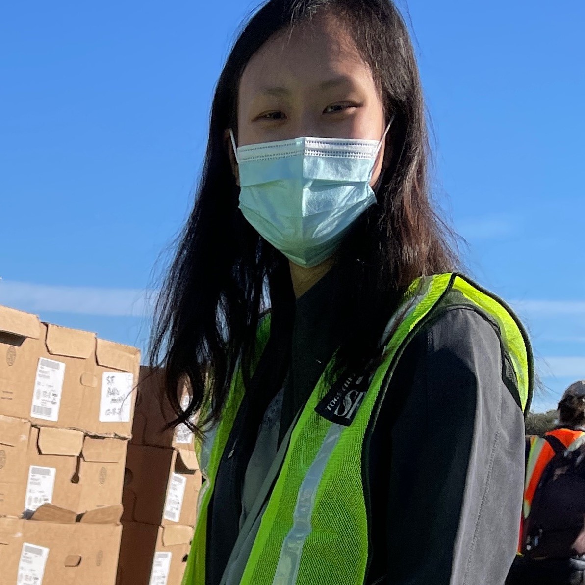 Mindy faces the camera smiling while wearing a blue facemask. She is wearing a yellow volunteer mesh jacket over her National Health Corps jacket.