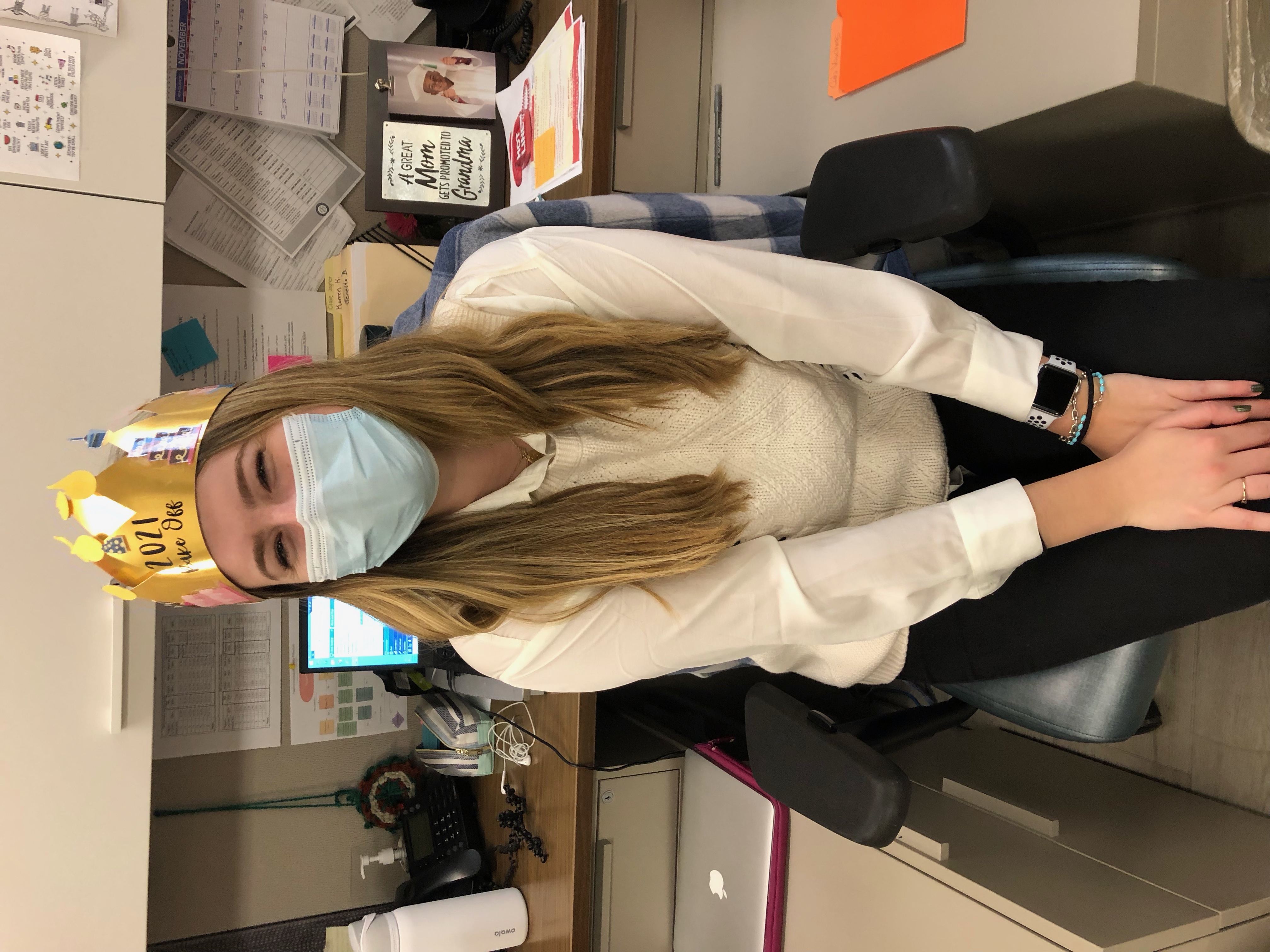 Claire sitting in a desk chair wearing a white sweater, a mask, and a paper crown