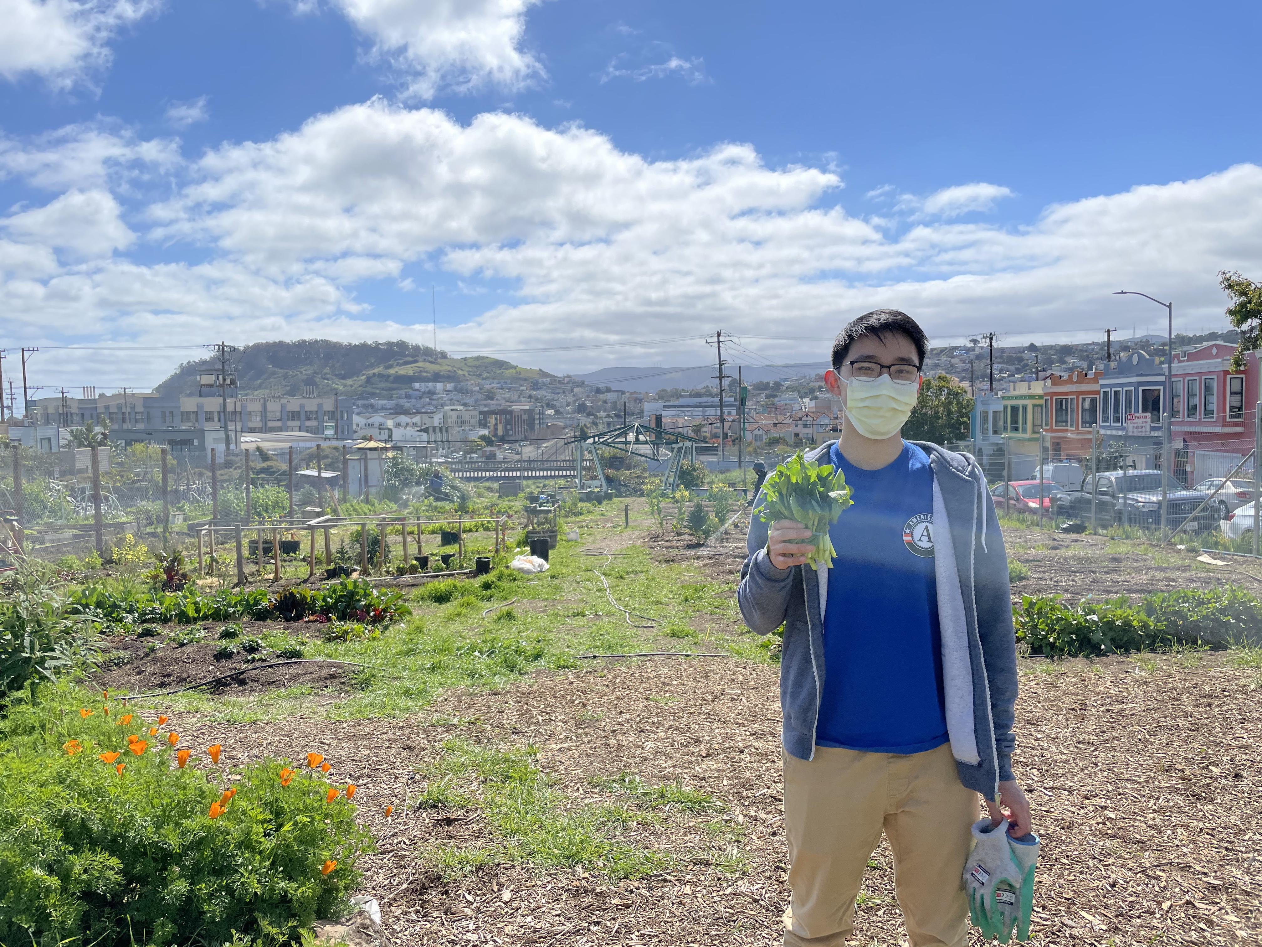 NHC SF Member Jacky Chu, wearing a sweater and their AmeriCorps shirt, holding a plant in their right hand with the Florence Fang Community Garden in the background. 
