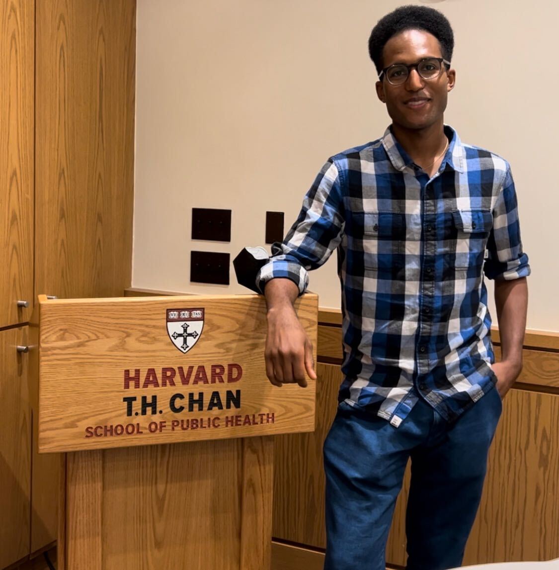 Spencer stands to the right of the frame, smiling at the camera. He is standing next to the podium showing the words "Harvard T.H. Chan School of Public Health"