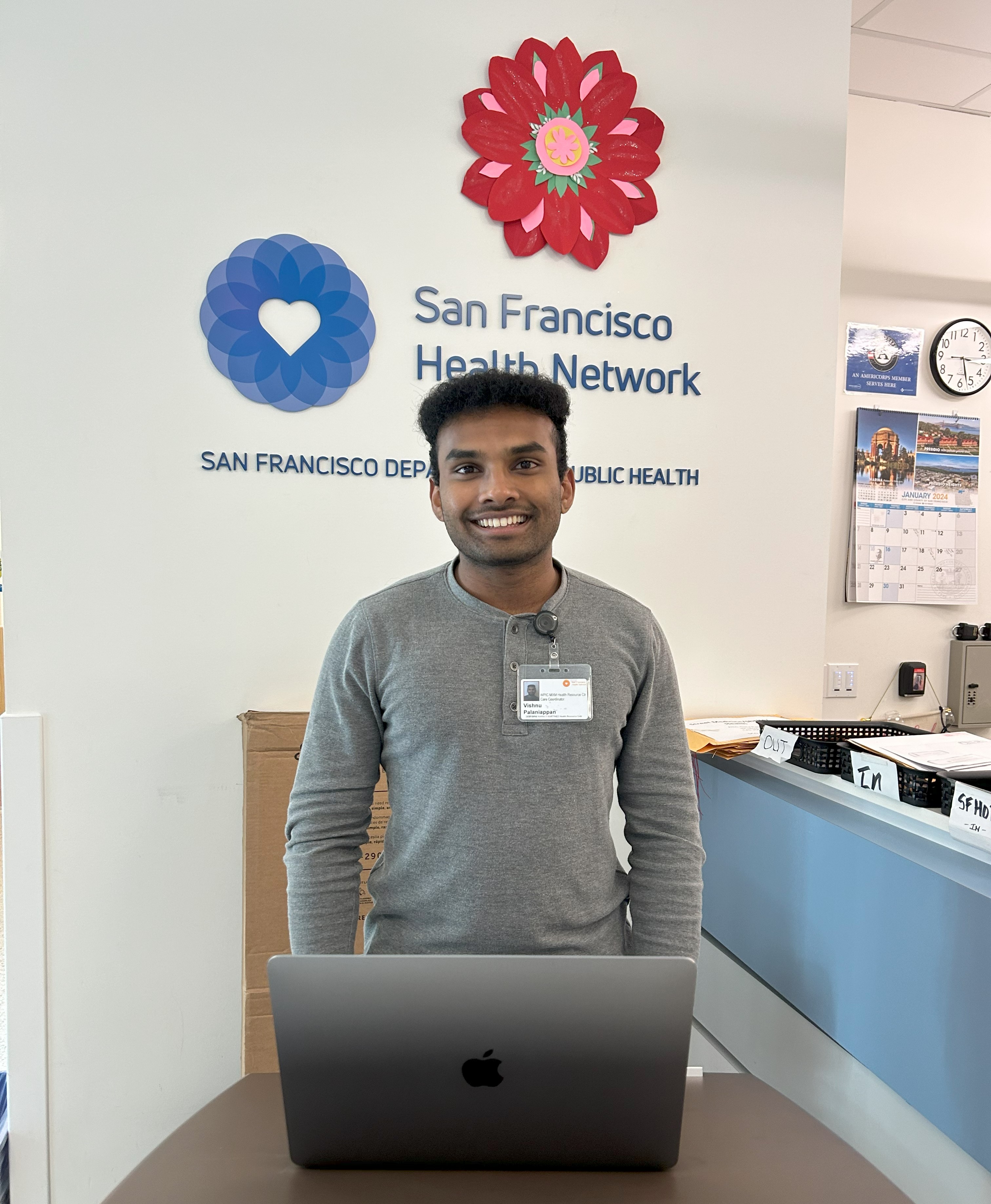 Vishnu is standing in the middle of the frame, facing the camera. He is standing behind a podium and has his laptop set on the podium in front of him. Behind him is the San Francisco Health Network logo. 