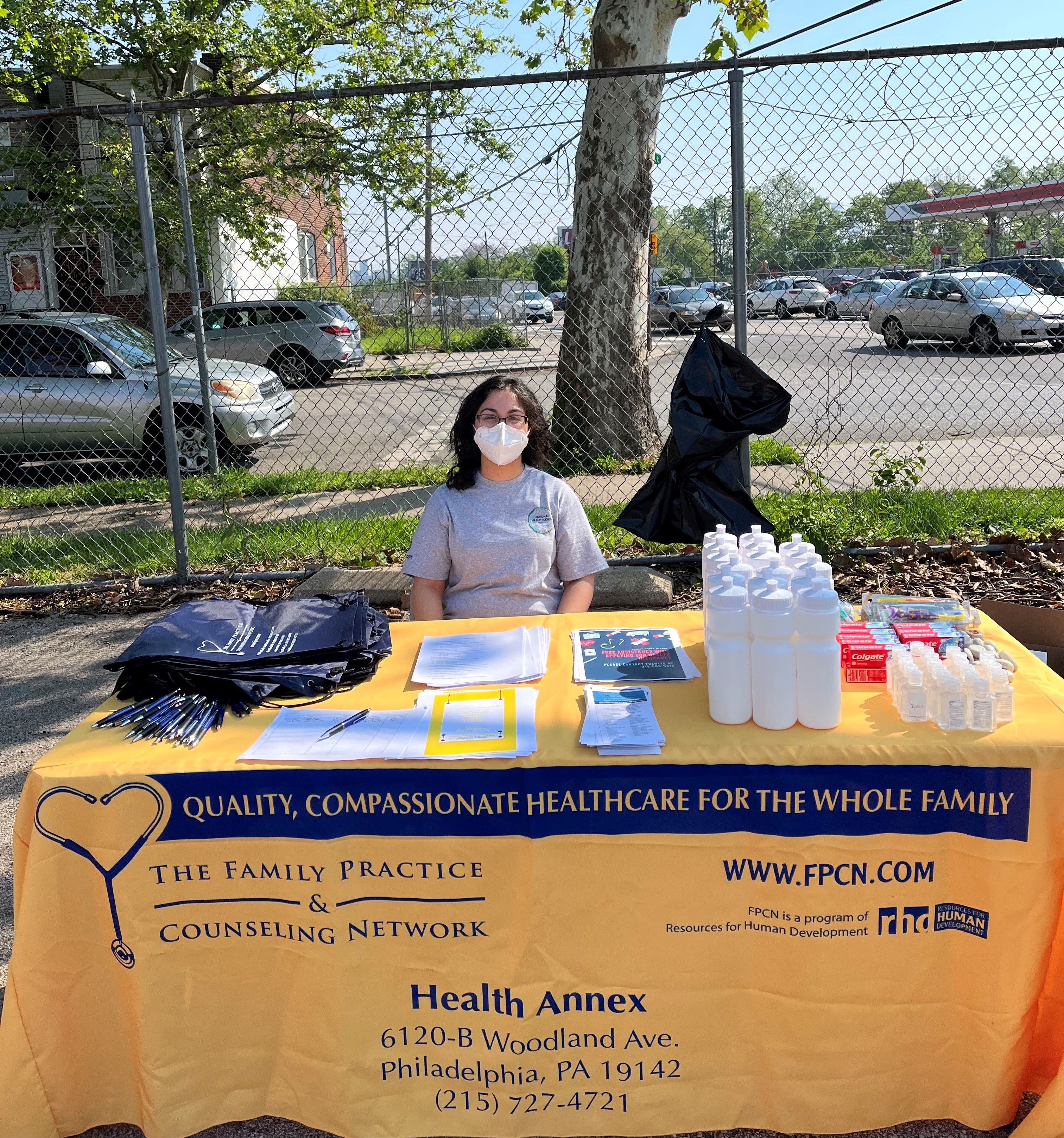 Amal sitting at a table covered in a yellow cloth advertising the Health Annex. On the table are bags, water bottles, and other giveaways for community members.