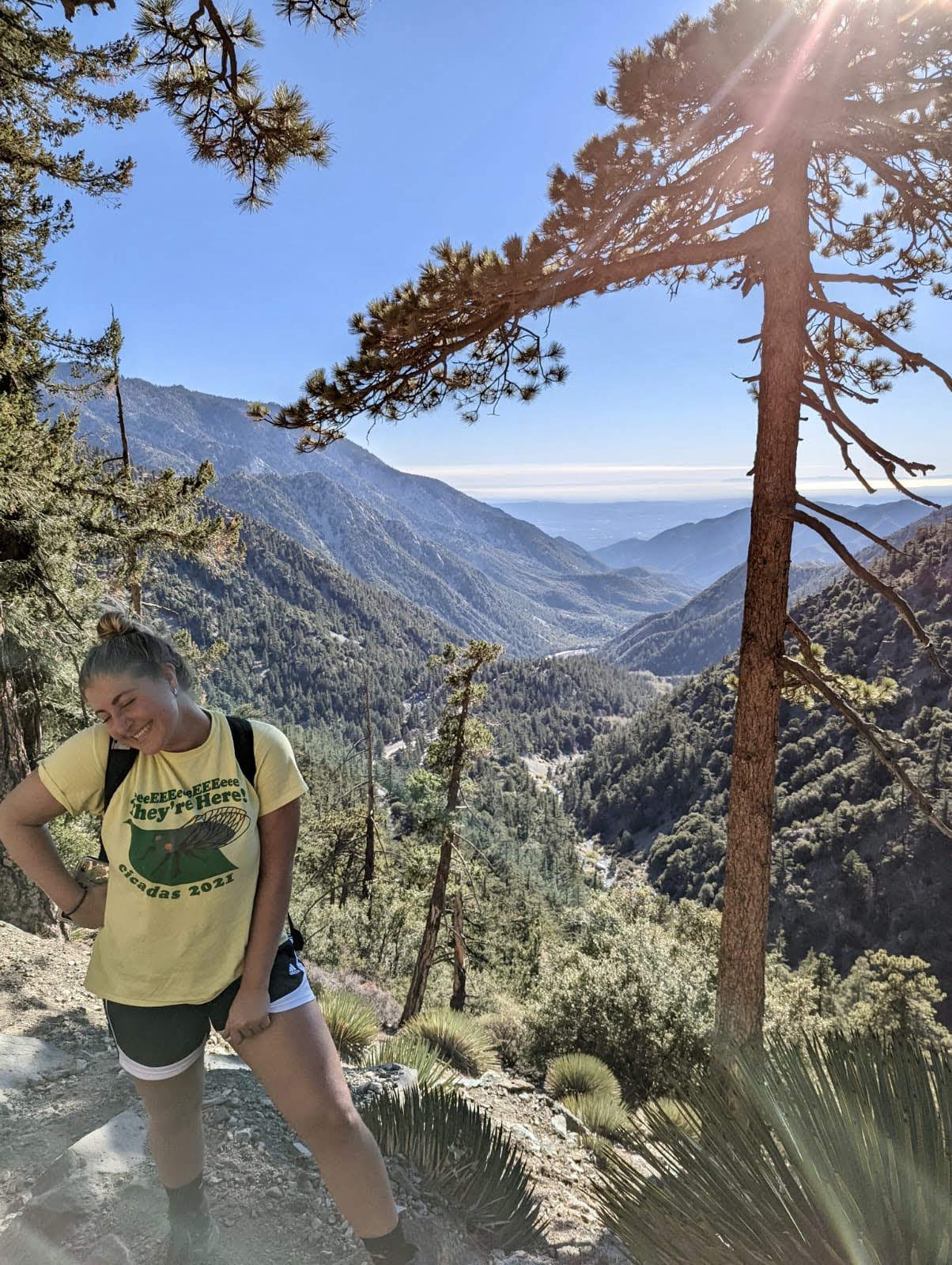 Sara in a yellow shirt and black shorts standing on a rocky ledge overlooking mountain covered in green trees