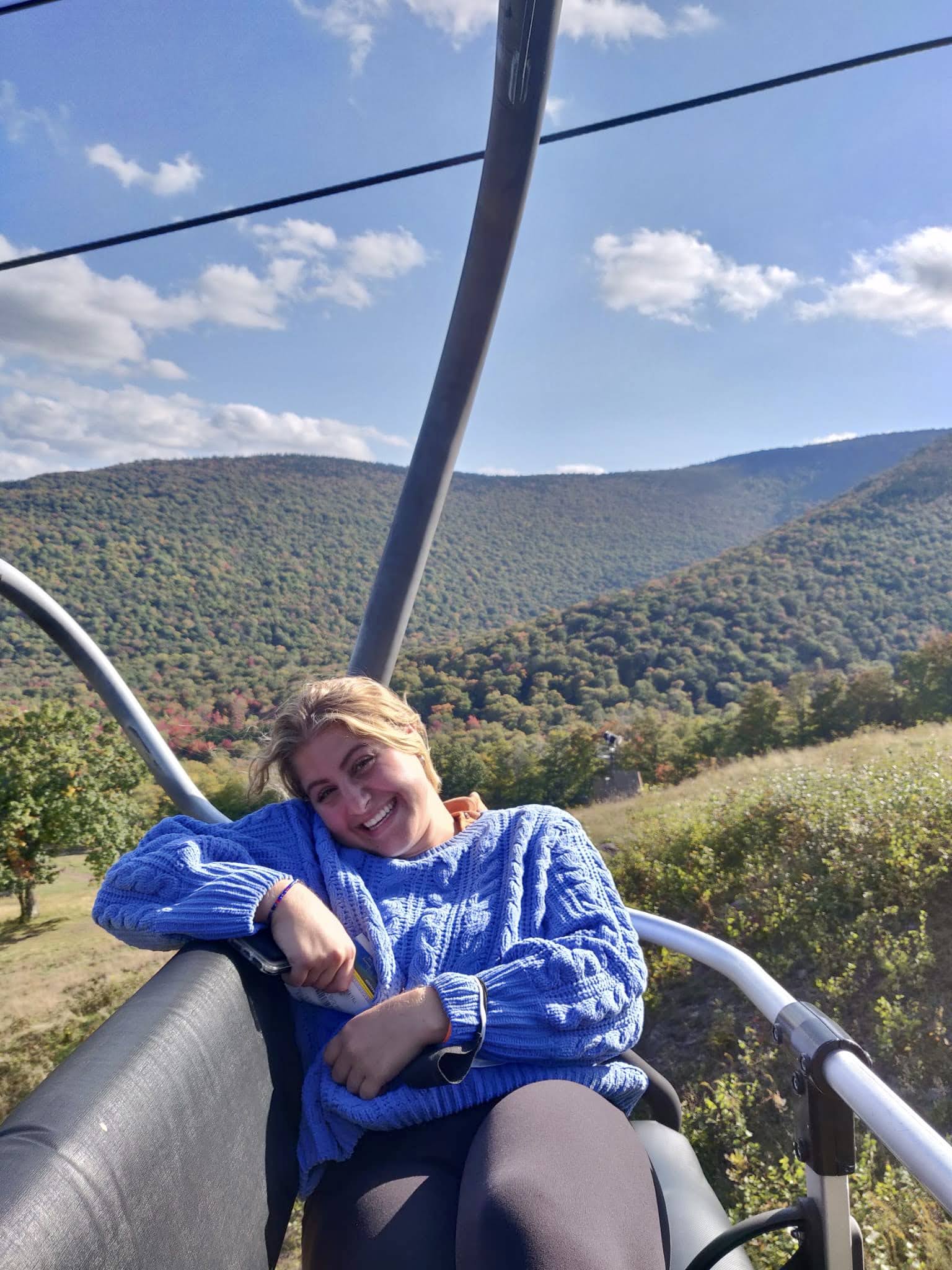 Sarah wearing a blue sweater sitting on a ski-lift during the summer, there are hills covered in green trees behind her.