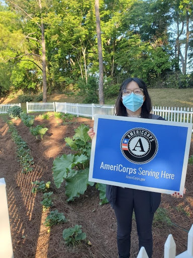 Jennifer standing in a community garden holding up a sign that reads "AmeriCorps Serving Here"