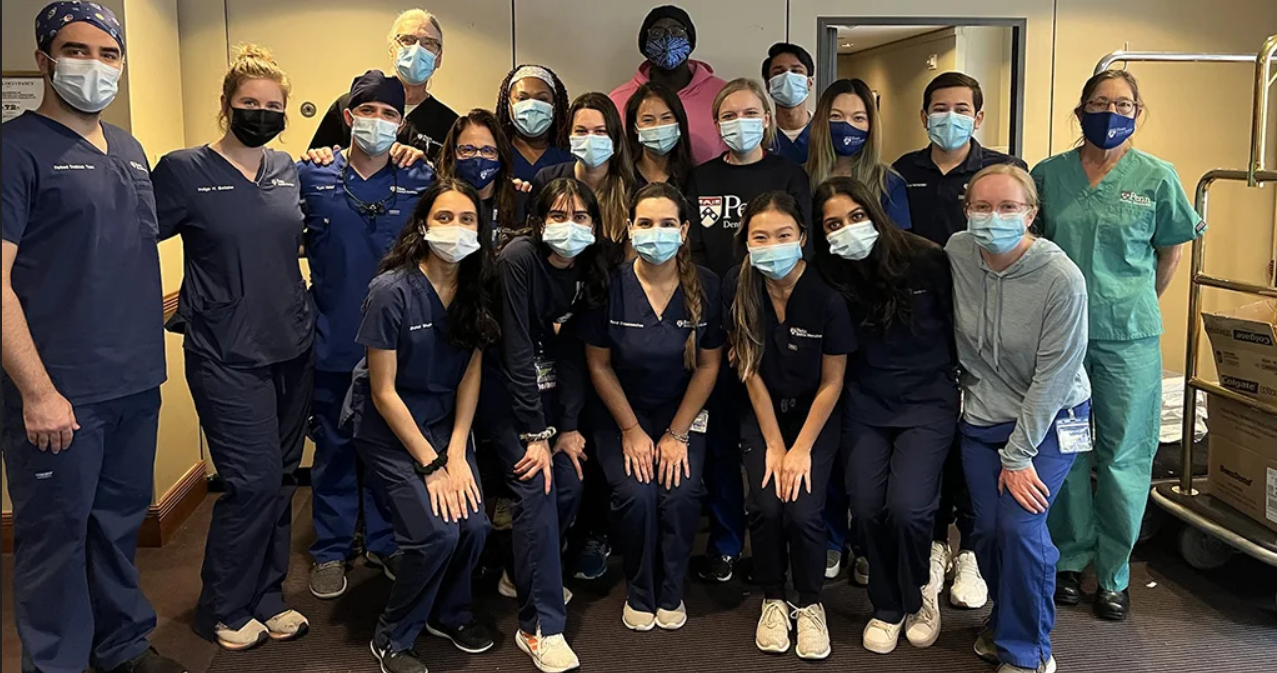 A group of people in blue scrubs and masks with one person in a pink hoodie in the back and one person in a gray hoodie on the right of the group. All are posed and looking at the camera, presumably smiling under their masks.
