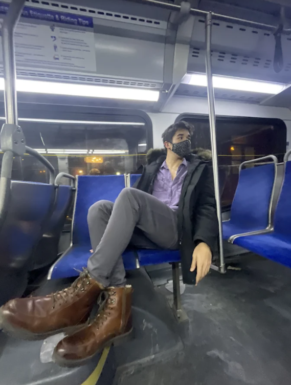Nathan sitting alone on a bus wearing am ask and looking back. His brown boots are in the foreground of the photo.